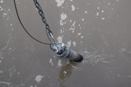 an ISE Max probe submerged in an aeration basin. The probe is a long black cylinder connected to a cord and a chain for security. 